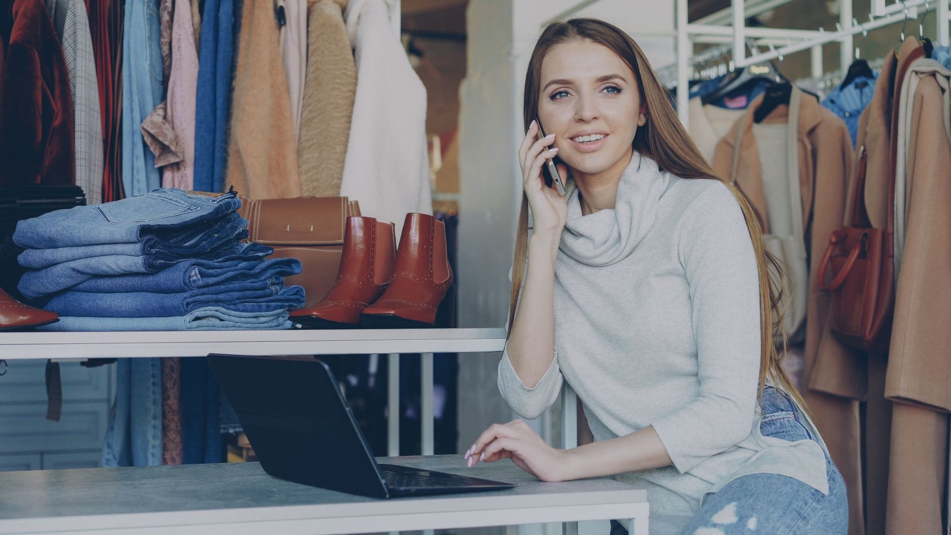 Sprzedawczyni w butiku rozmawia przez telefon i zarządza ofertami na laptopie